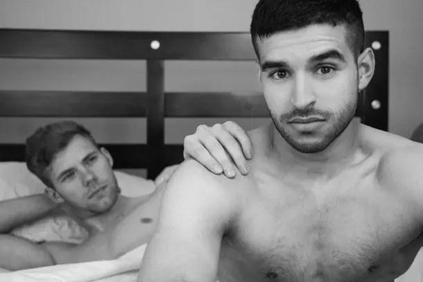 Two men lying on a bed, one gently resting a hand on the other’s shoulder, both appearing thoughtful symbolising intimacy, reflection, and the importance of support for gay relationships