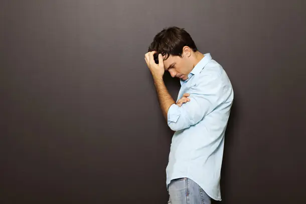 Man standing against a dark wall with head lowered and hand in his hair, appearing deep in thought symbolising emotional struggle and the possible need for support