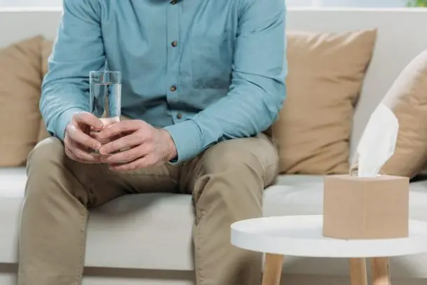 Person sitting on a sofa holding a glass of water, with a box of tissues on the table nearby, symbolising a safe, supportive counselling environment