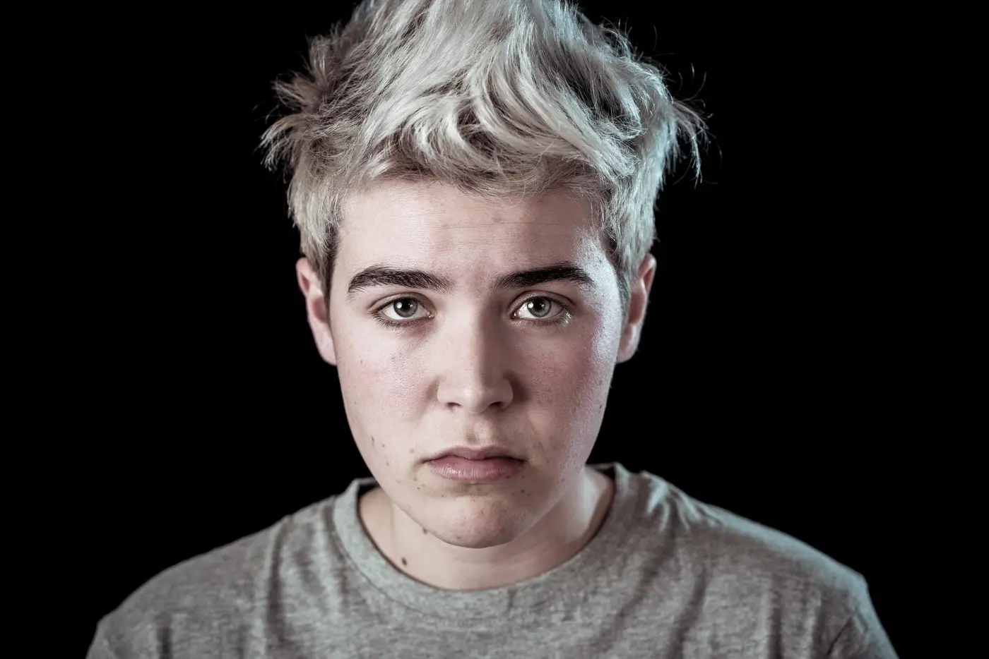 Young person with short, light hair looking directly at the camera with a thoughtful, slightly sad expression, symbolising a moment of vulnerability and a possible need for mental health support