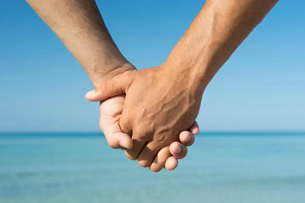 Close-up of two men holding hands by the sea, symbolising love, connection, and support for gay relationships