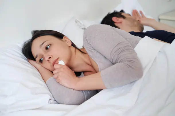 Woman lying in bed looking upset and holding a tissue while her partner sleeps beside her, symbolising emotional pain, loneliness, or relationship difficulties