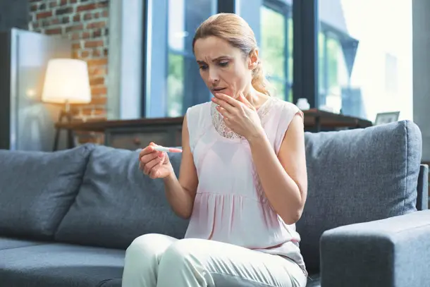 Woman sitting on a sofa looking concerned while holding a pregnancy test, reflecting the emotional impact and uncertainty that can come with infertility