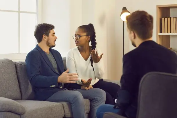 a distant view of a couple sitting on a sofa and talking with a therapist during a counselling session, expressing emotions in a safe and supportive environment.