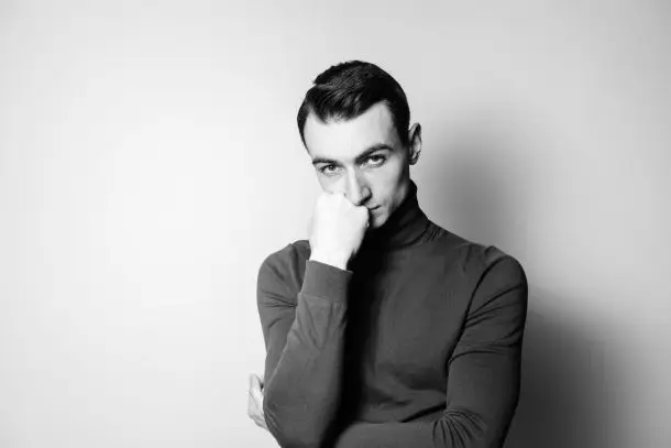 Black and white portrait of a man standing with one hand partly covering his mouth, looking thoughtful and pensive, symbolising reflection and the possible need for emotional support.