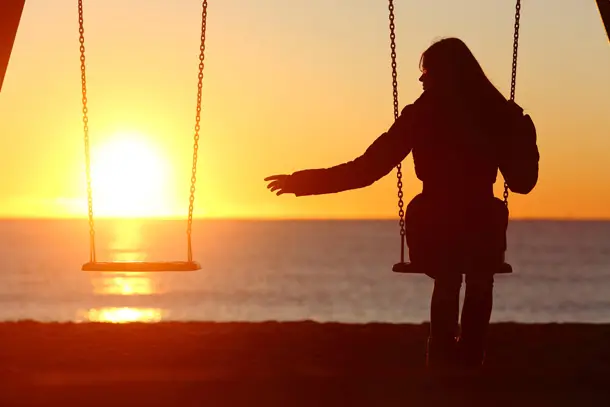 Silhouette of a person sitting alone on a swing at sunset, reaching toward an empty swing beside them symbolising grief, loss, and longing